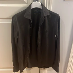 Svart skjorta från Polo Ralph Lauren - Säljer en stilren svart/grå skjorta från Polo Ralph Lauren. Skjortan har långa ärmar och knappar framtill. Den är broderad med den ikoniska logotypen på bröstet. Perfekt för en elegant look.