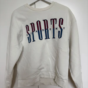 Vit sweatshirt från Gina Tricot - Säljer en vit sweatshirt från Gina Tricot i storlek S med texten 'SPORTS ACADEMY' i blått och rött på framsidan. Tröjan har långa ärmar och en rund halsringning. Säljer då den inte används. 