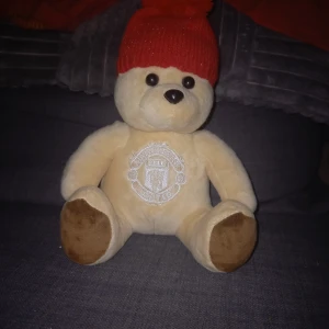 Manchester United Nalle Röd stickad mössa - Manchester United nalle med röd stickad mössa med en stor tofs på toppen. Perfekt för att hålla värmen under kalla dagar. Enkel och stilren design som passar till det mesta.