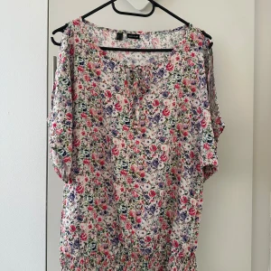 Blommig blus från Bodyflirt - Charmig blommig blus från Bodyflirt med korta ärmar och knytning framtill. Blusen har ett färgglatt mönster med rosa, lila och gröna blommor på en vit bakgrund. Perfekt för en somrig look!