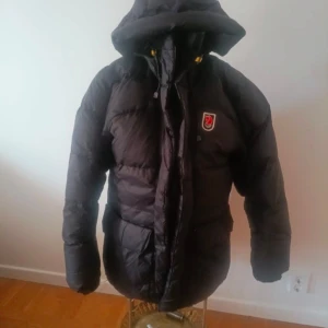 Svart dunjacka från Fjällräven - Säljer en svart dunjacka från Fjällräven med huva och dragkedja. Jackan har ett broderat märke på bröstet och långa ärmar. Perfekt för kalla dagar. jackan är storlek medium