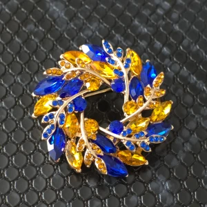 Blå och gul brosch - Elegant brosch i form av en krans med blå och gula stenar. Designen har en guldig bas som ger en lyxig känsla. Perfekt för att liva upp en outfit med sina klara färger och glittriga detaljer.