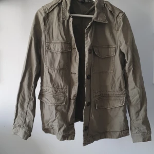 Grön jacka från H&M - Säljer en grön jacka från H&M med knappar framtill och flera praktiska fickor. Jackan har en klassisk krage, långa ärmar och dold dragsko i midjan på insidan. Ljus militärgrön skulle jag säga att färgen är! 100% bomull så superskön oavsett väder 💚