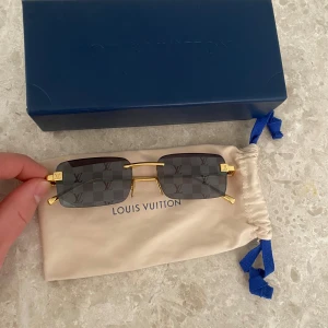 Solglasögon från Louis Vuitton - Snygga solglasögon från Louis Vuitton med rektangulära linser i grått och ett diskret rutmönster. Bågarna är i guldmetall med märkets logotyp på sidan. Perfekt accessoar för en stilren look.