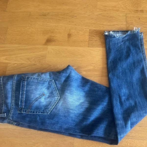 Dondup  - Säljer ett par blå jeans med slitna detaljer och fransiga benslut. Jeansen har en klassisk femficksdesign och en knappgylf. Perfekta för en avslappnad stil.