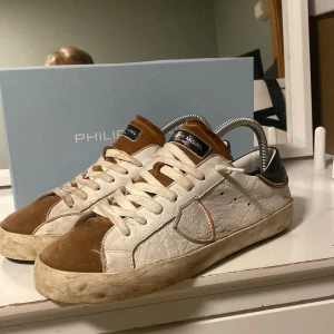 Vita och bruna sneakers från Philippe Model - Snygga sneakers från Philippe Model i vitt och brunt med svarta detaljer. Skorna har en klassisk design med snörning och en rund tå. Perfekta för en stilren look.