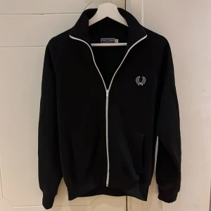 Svart jacka från Fred Perry - Snygg svart jacka från Fred Perry med vit dragkedja och broderad logga på bröstet. Perfekt för en stilren look. Jackan har långa ärmar och en klassisk krage. Storlek: XS. Hör av dig vid frågor ;)