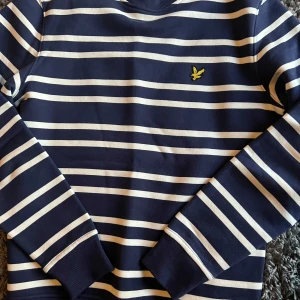 Randig tröja från Lyle & Scott - Säljer en stilren randig tröja från Lyle & Scott i marinblått och vitt. Tröjan har långa ärmar och ett broderat gult emblem på bröstet. Perfekt för en avslappnad look.