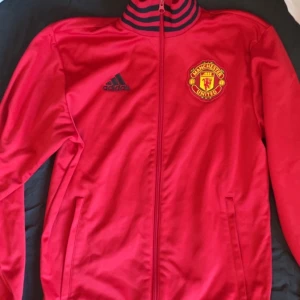 Röd Manchester United jacka från Adidas - Säljer en röd Manchester United jacka från Adidas med dragkedja och klubbens emblem på bröstet. Jackan har långa ärmar och en ribbad krage med svarta detaljer. Perfekt för fotbollsfans!