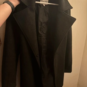 Svart kappa från NA-KD - Elegant svart kappa från NA-KD i storlek 36. Den har en klassisk design med knytbälte i midjan och långa ärmar. Perfekt för kyligare dagar med sin stilrena look, passar bra vår å höst - ”luddigt” material