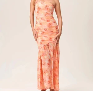 Adoore Blommig axelbandslös långklänning - Säljer en adoore axelbandslös långklänning med blommigt mönster i orange och rosa toner. Klänningen har en smickrande passform med rynkade detaljer och en elegant slits. Perfekt för sommarens festligheter! Helt ny från adoore, alla lappar kvar, endast testad bild 2 och 3 är mina egna bilder