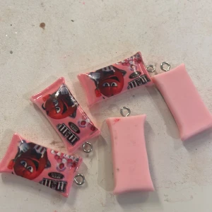 Rosa M&M's berlock - Charmiga rosa berlocker formade som M&M's-påsar med en röd karaktär på framsidan. Perfekta som accessoarer för att ge en lekfull touch till dina smycken eller nyckelringar. Säljer alla fem för 10kr