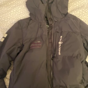 Svart vinterjacka från Sail Racing - Säljer en svart vinterjacka från Sail Racing med huva och dragkedja. Jackan har en logotyp på bröstet och en på ärmen. Perfekt för kalla dagar!