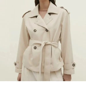 Beige trenchcoat med bälte - Snygg beige trenchcoat med knappar och knytbälte i midjan. Jackan har långa ärmar och en klassisk krage. Perfekt för en stilren look.