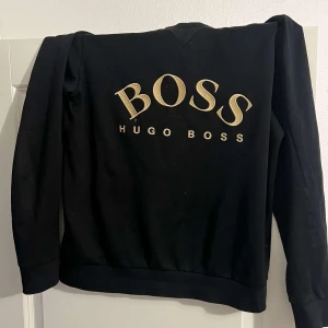 Hugo boss tröja - Snygg svart sweatshirt från Hugo Boss med stort guldigt BOSS-tryck på framsidan. Tröjan har långa ärmar och en klassisk rund halsringning. Perfekt för en stilren och avslappnad look.