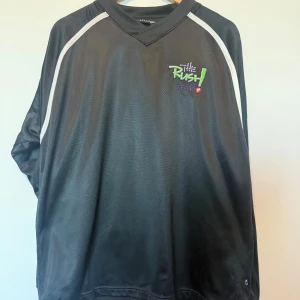 Svart tröja med tryck från The Rush Team - Säljer en svart tröja från The Rush Team med färgglatt tryck på framsidan. Tröjan har långa ärmar och vita detaljer längs axlarna och ryggen. Perfekt för en avslappnad stil.