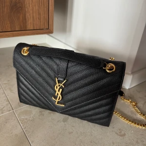 Svart quiltad axelväska Y*L - Elegant svart axelväska med quiltat mönster och guldfärgade detaljer. Väskan har en kedjerem och ett ikoniskt Y*L-emblem på framsidan. Perfekt för att ge en lyxig touch till din outfit.