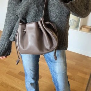 Brun axelväska i skinn - Snygg brun axelväska i skinn med en stilren design. Väskan har en praktisk dragsko för enkel öppning och stängning. Perfekt för att bära med sig det viktigaste med stil.