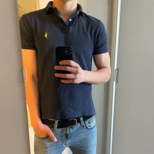 Mörkblå pikétröja från Polo Ralph Lauren - Säljer en klassisk mörkblå pikétröja från Ralph Lauren med den ikoniska loggan broderad i gult på bröstet. Tröjan har korta ärmar och en stilren krage med knappar. Perfekt för en avslappnad och stilren look. Skulle säga 9/10 skick eftersom defekter saknas men finns täcken på användning. Storlek S och är lite liten för mig som har M. Priset går att diskutera 