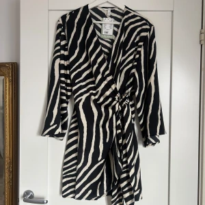 Zebramönstrad omlottklänning - Snygg zebramönstrad omlottklänning med trekvartsärmar och knytning i midjan. Perfekt för en trendig look med sin v-ringade design och svartvita ränder. Aldrig använd