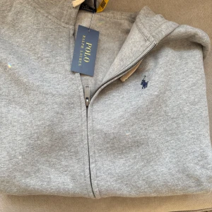 Grå hoodie från Polo Ralph Lauren - Säljer en stilren grå hoodie från Polo Ralph Lauren med dragkedja och det klassiska logotypbroderiet på bröstet. Perfekt för en avslappnad look. Den har en bekväm passform och är tillverkad i mjukt material.