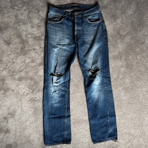 Blå jeans från Nudie Jeans - Säljer ett par blå jeans från Nudie Jeans med slitna detaljer på knäna och fransiga benslut. Jeansen har en klassisk femficksdesign och orange sömmar som ger en snygg kontrast.