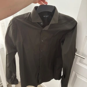 Olivgrön skjorta från Tiger of Sweden - Snygg olivgrön skjorta från Tiger of Sweden med klassisk krage och knappar framtill. Skjortan har långa ärmar och en stilren design som passar perfekt för en elegant look.