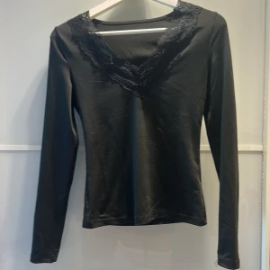 Svart långärmad topp med spetsdetaljer - Snygg svart långärmad topp med v-ringad spetsdetalj runt halsen. Perfekt för en stilren look. Passar bra till både jeans och kjol.