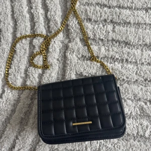 Svart quiltad axelväska med guldkedja - Snygg svart quiltad axelväska med guldkedja. Perfekt för att ge en elegant touch till din outfit. Väskan har en stilren design med en liten guldfärgad detalj på framsidan.