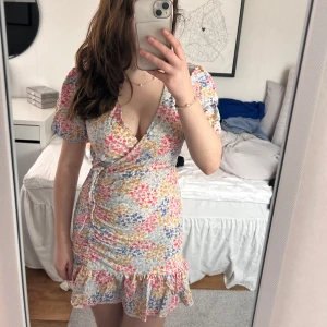 Blommig omlottklänning - Charmig kort blommig omlottklänning med volangdetaljer. Klänningen har en v-ringad design och korta ärmar, perfekt för sommarens varma dagar. Den har en knytning i midjan som ger en smickrande passform. Passar perfekt till sommaren och fantastisk till midsommar 💕