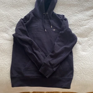Svart hoodie från GANT - Säljer en stilren svart hoodie från GANT i storlek M. Den har en klassisk design med justerbar huva och en praktisk magficka. Perfekt för en avslappnad look.
