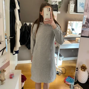 Grå långärmad hoodieklänning - Mysig grå långärmad hoodieklänning med en avslappnad passform. Klänningen har en stor ficka framtill och en bekväm huva. Perfekt för en chill dag hemma eller en avslappnad utflykt.