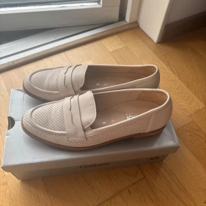 Beige loafers från Gabor - Snygga beige loafers från Gabor med perforerat mönster och klassisk design. Skorna har en rund tå och är tillverkade i skinn, vilket ger en elegant och tidlös look. Perfekta för en stilren vardagsstil.