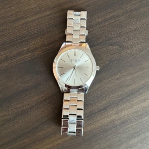 Silverfärgad klocka från Jet Set - Snygg och stilren klocka från Jet Set i silverfärg. Klockan har en metallarmband och en rund urtavla med minimalistisk design. Perfekt för att ge en elegant touch till din outfit.