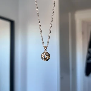 Guldhalsband med knutdesign - Elegant guldhalsband med en unik knutdesign som hänge. Kedjan är tunn och smidig, vilket gör den perfekt för att bära varje dag eller vid speciella tillfällen. Hänget har en intrikat flätad stil som ger en sofistikerad touch. Önskar du veta exakta mått? Tveka inte att höra av dig!