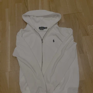 Vit hoodie från Ralph Lauren - Säljer en stilren vit hoodie från Ralph Lauren med dragkedja och det klassiska logotypbroderiet på bröstet. Hoodien har långa ärmar och en bekväm passform. Perfekt för en avslappnad look. Pris kan ändras på snabb affär 🙌🏽
