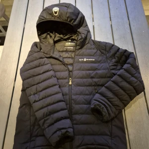 Svart jacka från Sail Racing - Säljer en svart dunjacka från Sail Racing med huva och dragkedja. Jackan är quiltad och har en stilren design med logotyp på bröstet. Perfekt de vardagar som väntar oss.