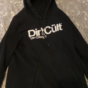 Svart hoodie från DirtCült - Säljer en svart hoodie från DirtCült med texten 'San Diego' tryckt i vitt på framsidan. Den har en klassisk design med dragsko i huvan och en stor magficka. Perfekt för en avslappnad stil.