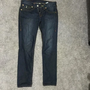 Mörkblå jeans från True Religion (dm för mer bilder) - Snygga mörkblå jeans från True Religion med ikonisk broderad design på bakfickorna. De har en klassisk femficksmodell och knappar i silverfärg. Perfekta för en stilren look.