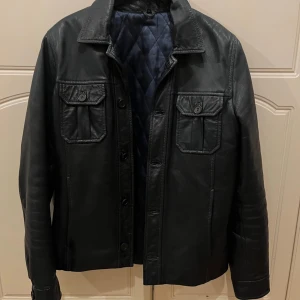 Mörkblå skinnjacka - Säljer en stilren mörkblå skinnjacka med knappar och två bröstfickor. Jackan har en quiltad insida för extra komfort och värme. Perfekt för en cool look när det är lite kyligare ute. Passar bra till jeans och sneakers. Bra skick, nypris ca 1500kr