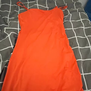 Neon Orange klänning från H&M Divided - Snygg orange klänning från H&M Divided med smala axelband och rynkade detaljer på sidan. Perfekt för en färgglad look. Klänningen är i en kort modell och har en figurnära passform. Den e inte så kort til knäna är den men knappt använt kanske 3 gånger.
