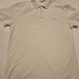 Vit pikétröja från Lacoste - Vit pikétröja från Lacoste med korta ärmar och den ikoniska krokodilloggan på bröstet. 