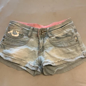 Lågmidjade Jeansshorts - Snygga jeansshorts från okänt märke. Detaljer på bakfickorna. De har en ljusblå tvätt och rosa insida. Shortsen har dubbla knappar och nitar vid fickorna för extra stil. Perfekta för sommardagar! Det kan vara gamla true religon