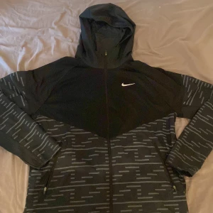 Svart och grå mönstrad windbraker från Nike - Säljer en snygg svart och grå windbraker från Nike med ett unikt mönster. Den har en dragkedja framtill och en praktisk huva. Perfekt för en sportig look eller avslappnade dagar.