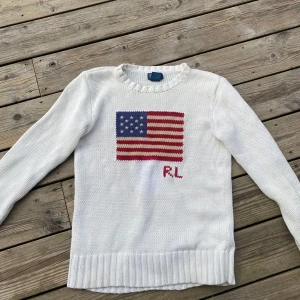 Vit stickad tröja med usa flagga från Polo Ralph Lauren - Säljer en vit stickad tröja från Polo Ralph Lauren med en amerikansk flagga och RL-broderi på framsidan. Tröjan har långa ärmar och ribbstickade detaljer vid hals, ärmslut och nederkant. Perfekt för en stilren och avslappnad look.