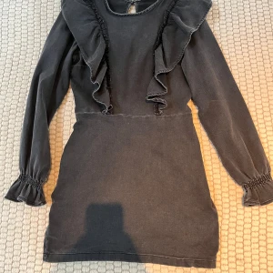 Svart klänning med volangdetaljer - Snygg svart klänning med volangdetaljer framtill och långa ärmar. Perfekt för en stilren look. Klänningen har en smickrande passform och elastiska ärmslut för extra komfort.