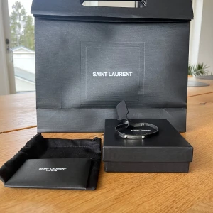 Svart armband från Saint Laurent - Elegant svart armband från Saint Laurent med en stilren design. Armbandet har en metallplatta med ingraverad logotyp och stängs med ett diskret spänne. Perfekt för att ge en lyxig touch till din outfit. 