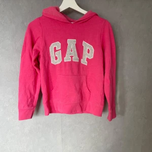 Rosa hoodie från GAP Kids - Säljer en snygg rosa hoodie från GAP Kids med stor logga på framsidan. Tröjan har en klassisk känguruficka och en mysig huva. Perfekt för en avslappnad stil.