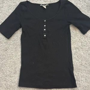 Svart ribbad topp med knappar - Säljer en stilren svart ribbad topp med korta ärmar och knappar framtill. Perfekt för en enkel och snygg look. Passar bra till både jeans och kjol.