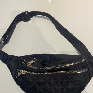 Svart quiltad midjeväska - Snygg svart quiltad midjeväska med dragkedjor. Perfekt för att hålla dina saker säkra och nära till hands. Justerbart band för bekväm passform.